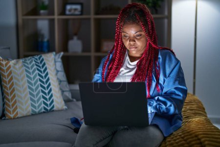 Foto de Mujer afroamericana con cabello trenzado usando computadora portátil en la noche relajado con expresión seria en la cara. simple y natural mirando a la cámara. - Imagen libre de derechos