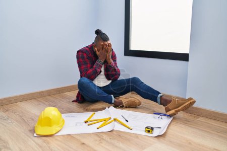 Foto de Hombre afroamericano sentado en el suelo en una casa nueva mirando planos con expresión triste cubriendo la cara con las manos mientras llora. concepto de depresión. - Imagen libre de derechos