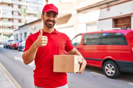Photo for Young hispanic man with beard wearing delivery uniform and cap holding box smiling happy and positive, thumb up doing excellent and approval sign - Royalty Free Image