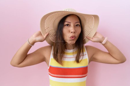 Téléchargez les photos : Femme chinoise du Moyen Âge portant un chapeau d'été sur fond rose regardant la caméra souffler un baiser étant belle et sexy. expression d'amour. - en image libre de droit