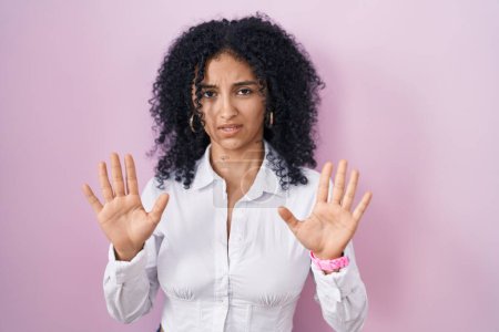 Foto de Mujer hispana con el pelo rizado de pie sobre fondo rosa alejando las palmas de las manos mostrando rechazo y negación con miedo y expresión repugnante. parada y prohibido. - Imagen libre de derechos