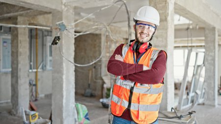 Téléchargez les photos : Jeune homme hispanique constructeur souriant confiant debout avec les bras croisés geste sur le chantier de construction - en image libre de droit