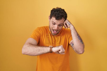 Téléchargez les photos : Jeune homme hispanique debout sur fond jaune regardant le temps de la montre inquiet, peur de se faire tard - en image libre de droit
