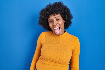 Foto de Mujer negra con el pelo rizado de pie sobre fondo azul sacando la lengua feliz con expresión divertida. concepto de emoción. - Imagen libre de derechos
