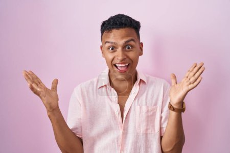 Foto de Joven hombre hispano de pie sobre fondo rosa celebrando loco y sorprendido por el éxito con los brazos levantados y los ojos abiertos gritando emocionado. concepto ganador - Imagen libre de derechos