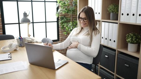 Foto de Trabajadora de negocios embarazada joven usando la escritura portátil en el cuaderno en la oficina - Imagen libre de derechos