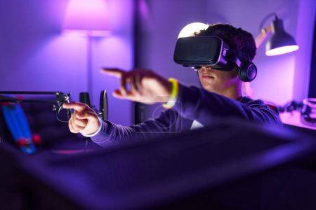 Téléchargez les photos : Jeune adolescent hispanique streamer jouer à un jeu vidéo en utilisant des lunettes de réalité virtuelle à la salle de jeu - en image libre de droit