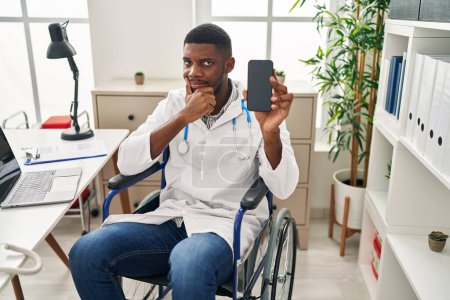 Foto de Hombre médico afroamericano sentado en silla de ruedas sosteniendo el teléfono inteligente cara seria pensando en la pregunta con la mano en la barbilla, reflexivo acerca de la idea confusa - Imagen libre de derechos