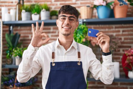 Téléchargez les photos : Jeune homme hispanique travaillant à la boutique de fleuriste tenant carte de crédit faisant signe ok avec les doigts, sourire geste amical excellent symbole - en image libre de droit