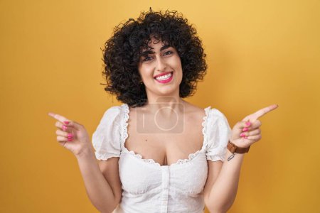 Foto de Mujer morena joven con el pelo rizado de pie sobre el fondo amarillo sonriendo confiado señalando con los dedos a diferentes direcciones. espacio de copia para publicidad - Imagen libre de derechos