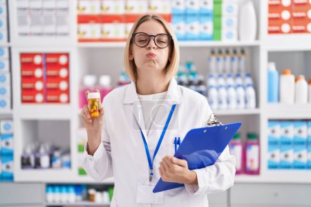 Téléchargez les photos : Jeune femme caucasienne travaillant à la pharmacie drugstore tenant des pilules gonflant les joues avec un visage drôle. bouche gonflée d'air, attrapant l'air. - en image libre de droit