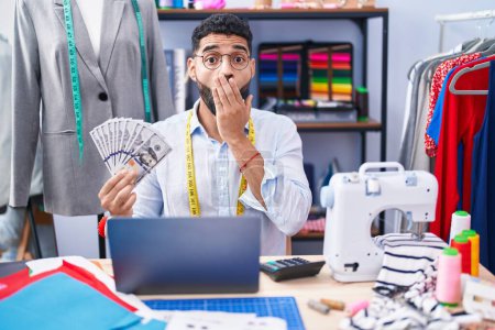 Photo for Hispanic man with beard dressmaker designer holding dollars covering mouth with hand, shocked and afraid for mistake. surprised expression - Royalty Free Image
