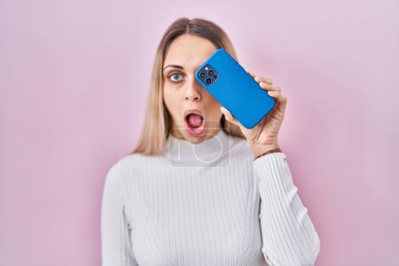 Foto de Mujer rubia joven sosteniendo teléfono inteligente que cubre el ojo asustado y sorprendido con la boca abierta para sorpresa, cara de incredulidad - Imagen libre de derechos