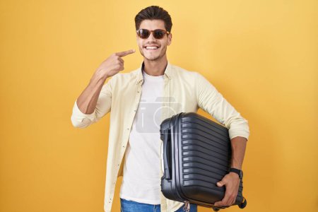 Téléchargez les photos : Jeune homme hispanique tenant une valise allant en vacances d'été souriant joyeuse montrant et pointant du doigt les dents et la bouche. concept de santé dentaire. - en image libre de droit