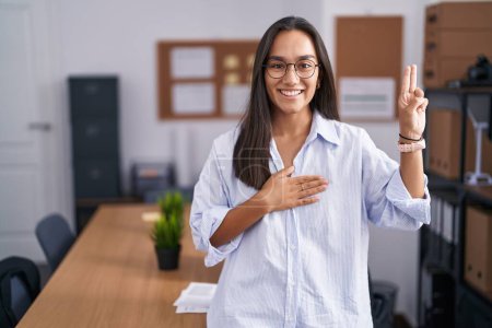 Foto de Mujer hispana joven en la oficina sonriendo jurando con la mano en el pecho y los dedos hacia arriba, haciendo un juramento de lealtad promesa - Imagen libre de derechos