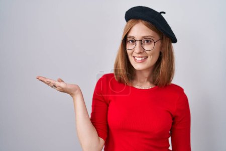 Téléchargez les photos : Jeune rousse femme debout portant des lunettes et béret souriant gai présentation et pointant avec la paume de la main regardant la caméra. - en image libre de droit