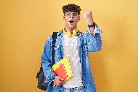 Photo for Hispanic teenager wearing student backpack and holding books angry and mad raising fist frustrated and furious while shouting with anger. rage and aggressive concept. - Royalty Free Image