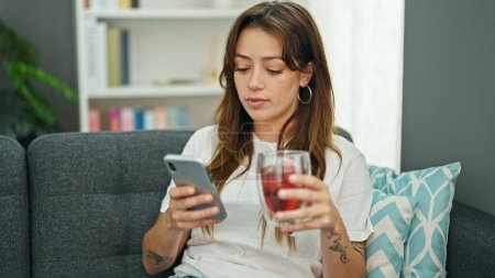 Téléchargez les photos : Jeune belle femme hispanique boire du thé à l'aide d'un smartphone à la maison - en image libre de droit