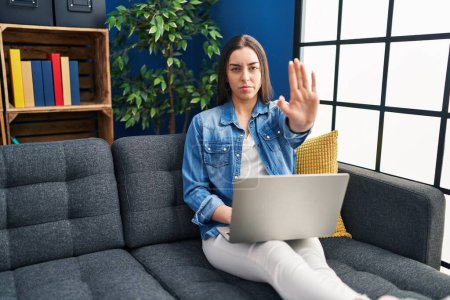 Foto de Mujer hispana usando laptop en casa con la mano abierta haciendo stop sign con expresión seria y segura, gesto de defensa - Imagen libre de derechos