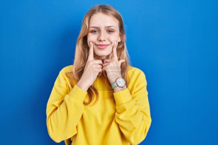 Photo for Young caucasian woman standing over blue background smiling with open mouth, fingers pointing and forcing cheerful smile - Royalty Free Image