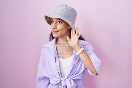 Téléchargez les photos : Jeune femme hispanique debout sur fond rose portant un chapeau souriant avec la main sur l'oreille en écoutant une rumeur ou des commérages. concept de surdité. - en image libre de droit