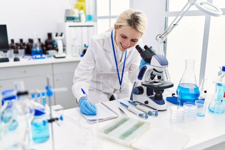 Téléchargez les photos : Jeune femme blonde scientifique utilisant un microscope prenant des notes au laboratoire - en image libre de droit