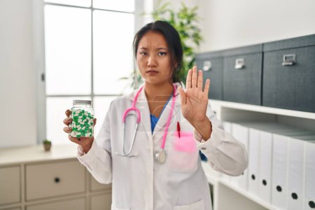 Photo for Young asian doctor woman holding pills with open hand doing stop sign with serious and confident expression, defense gesture - Royalty Free Image