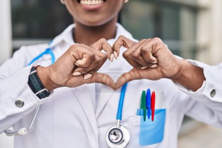 Téléchargez les photos : Femme afro-américaine médecin souriant confiant faire geste de coeur avec les mains à l'hôpital - en image libre de droit