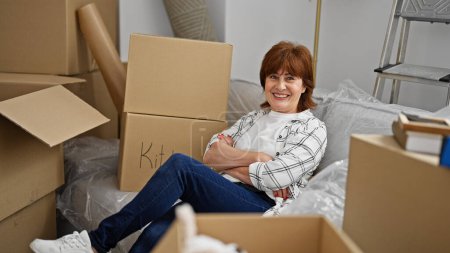 Téléchargez les photos : Femme d'âge moyen souriant confiant assis sur le canapé avec les bras croisés geste à la nouvelle maison - en image libre de droit