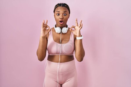 Photo for African american woman with braids wearing sportswear and headphones looking surprised and shocked doing ok approval symbol with fingers. crazy expression - Royalty Free Image