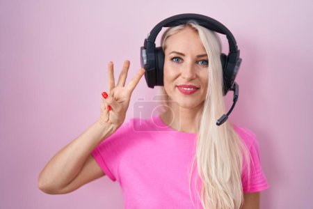 Téléchargez les photos : Femme caucasienne écoutant de la musique à l'aide d'écouteurs montrant et pointant vers le haut avec les doigts numéro trois tout en souriant confiant et heureux. - en image libre de droit