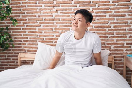 Téléchargez les photos : Jeune homme chinois souriant confiant assis sur le lit dans la chambre - en image libre de droit