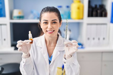 Foto de Mujer joven haciendo extracción de aceite de hierba en el laboratorio sonriendo con una sonrisa feliz y fresca en la cara. mostrando dientes. - Imagen libre de derechos