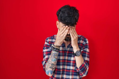 Photo for Young hispanic man with beard standing over red background rubbing eyes for fatigue and headache, sleepy and tired expression. vision problem - Royalty Free Image