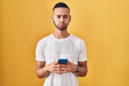 Téléchargez les photos : Jeune homme hispanique à l'aide d'un smartphone tapant message joues gonflées avec drôle de visage. bouche gonflée d'air, attrapant l'air. - en image libre de droit