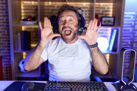 Foto de Hombre de mediana edad con barba jugando videojuegos con auriculares sonriendo alegre jugando a echar un vistazo a un boo con las manos mostrando la cara. sorprendido y salido - Imagen libre de derechos