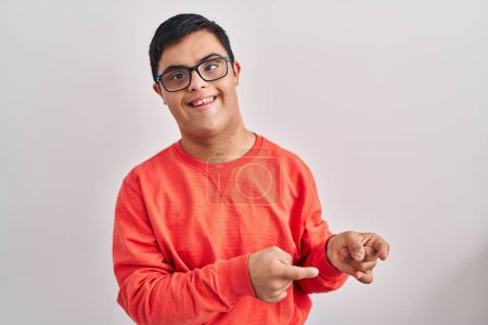 Foto de Joven hombre hispano con síndrome de Down de pie sobre fondo blanco sonriendo y mirando a la cámara apuntando con dos manos y dedos hacia un lado. - Imagen libre de derechos