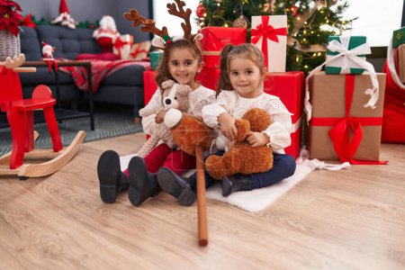 Foto de Dos niños sosteniendo oso de peluche sentado en el suelo por el árbol de Navidad en casa - Imagen libre de derechos