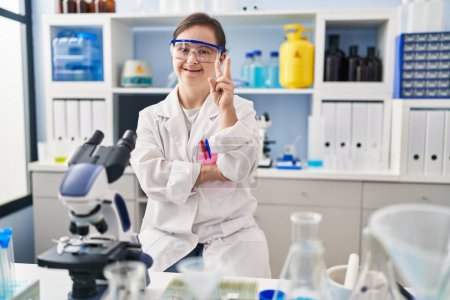 Photo for Hispanic girl with down syndrome working at scientist laboratory smiling with happy face winking at the camera doing victory sign with fingers. number two. - Royalty Free Image