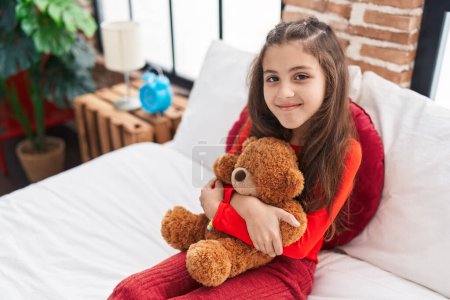 Photo for Adorable hispanic girl hugging teddy bear sitting on bed at bedroom - Royalty Free Image