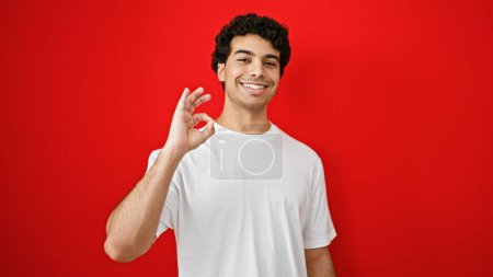 Foto de Joven latino haciendo buen gesto sonriendo sobre fondo rojo aislado - Imagen libre de derechos