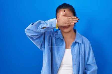 Photo for Asian young woman standing over blue background covering eyes with hand, looking serious and sad. sightless, hiding and rejection concept - Royalty Free Image