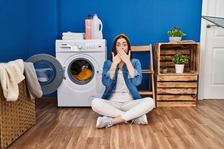 Photo for Hispanic woman doing laundry sitting on the floor shocked covering mouth with hands for mistake. secret concept. - Royalty Free Image
