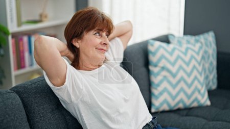 Téléchargez les photos : Femme d'âge moyen détendue avec les mains sur la tête assise sur le canapé maison - en image libre de droit