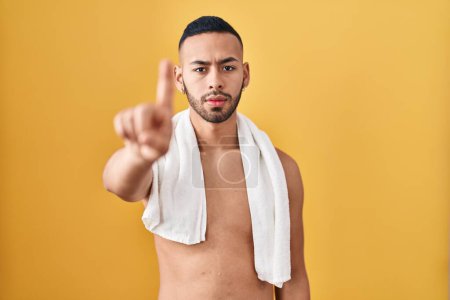 Téléchargez les photos : Jeune homme hispanique debout torse nu avec serviette pointant le doigt vers le haut et expression en colère, ne montrant aucun geste - en image libre de droit