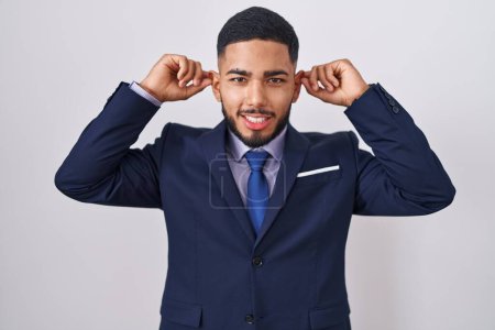 Foto de Joven hombre hispano con traje de negocios y corbata sonriente tirando de las orejas con los dedos, gesto divertido. problema de audición - Imagen libre de derechos