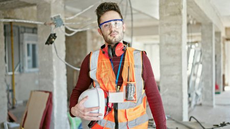 Téléchargez les photos : Jeune homme hispanique constructeur debout avec le visage sérieux tenant le casque dur sur le chantier de construction - en image libre de droit
