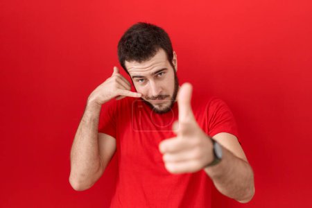 Foto de Un joven hispano vistiendo una camiseta roja casual sonriendo hablando por teléfono y señalándote. Llámame.. - Imagen libre de derechos
