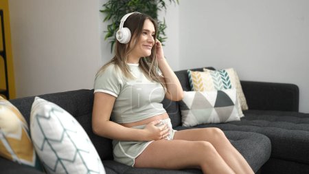 Foto de Mujer embarazada joven escuchando música tocando el vientre en casa - Imagen libre de derechos