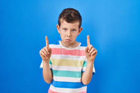Photo for Young caucasian kid standing over blue background pointing up looking sad and upset, indicating direction with fingers, unhappy and depressed. - Royalty Free Image